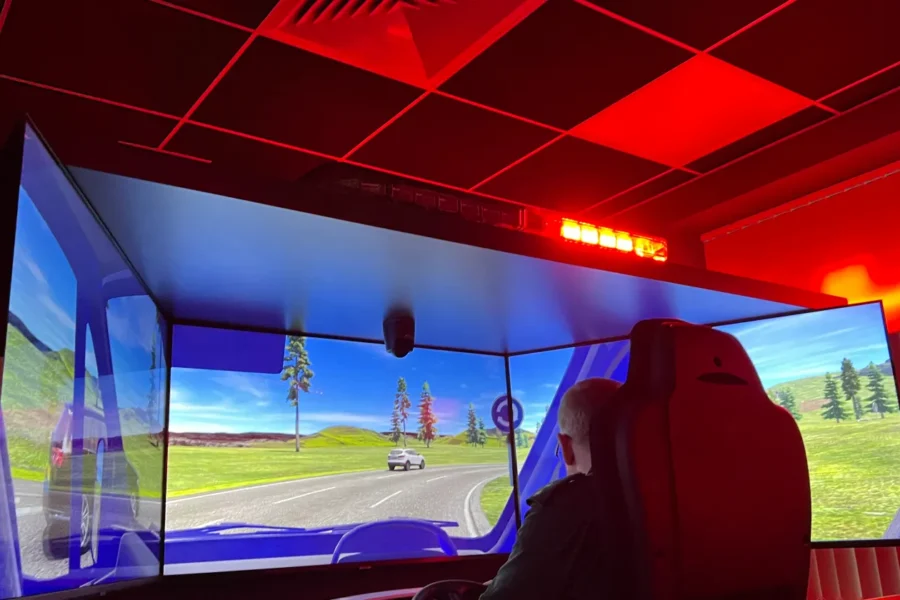 a man sat on a chair at a computer desk, 3 computer screens showing a virtual driving simulation