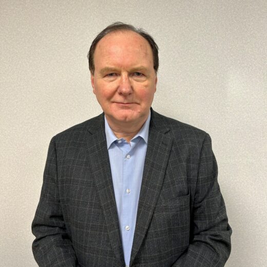 a photo of a man in a blue shirt, dark suit jacket in front of a grey background