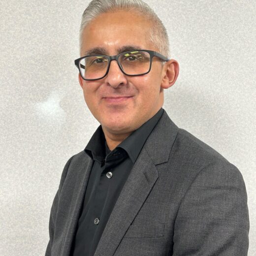 a man in a black shirt and suit jacket. Wearing glasses, in front of a white show