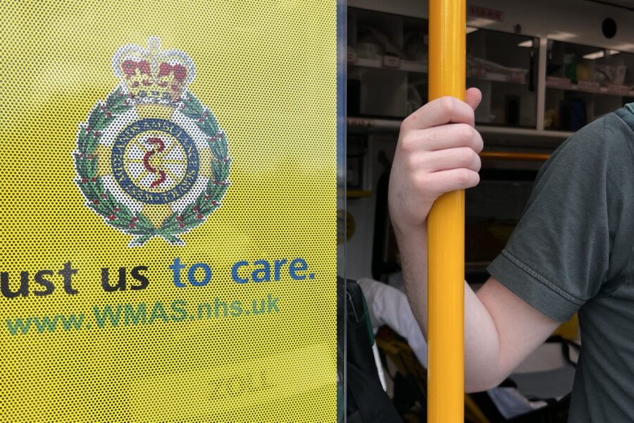 Paramedic's hand holding yellow grab bar on back of ambulance with 'Trust us to care WMAS' logo on racking wall